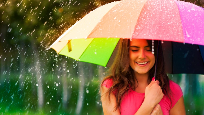 Woman smiling under a colorful umbrella in the rain, representing financial protection and a rainy day savings fund.